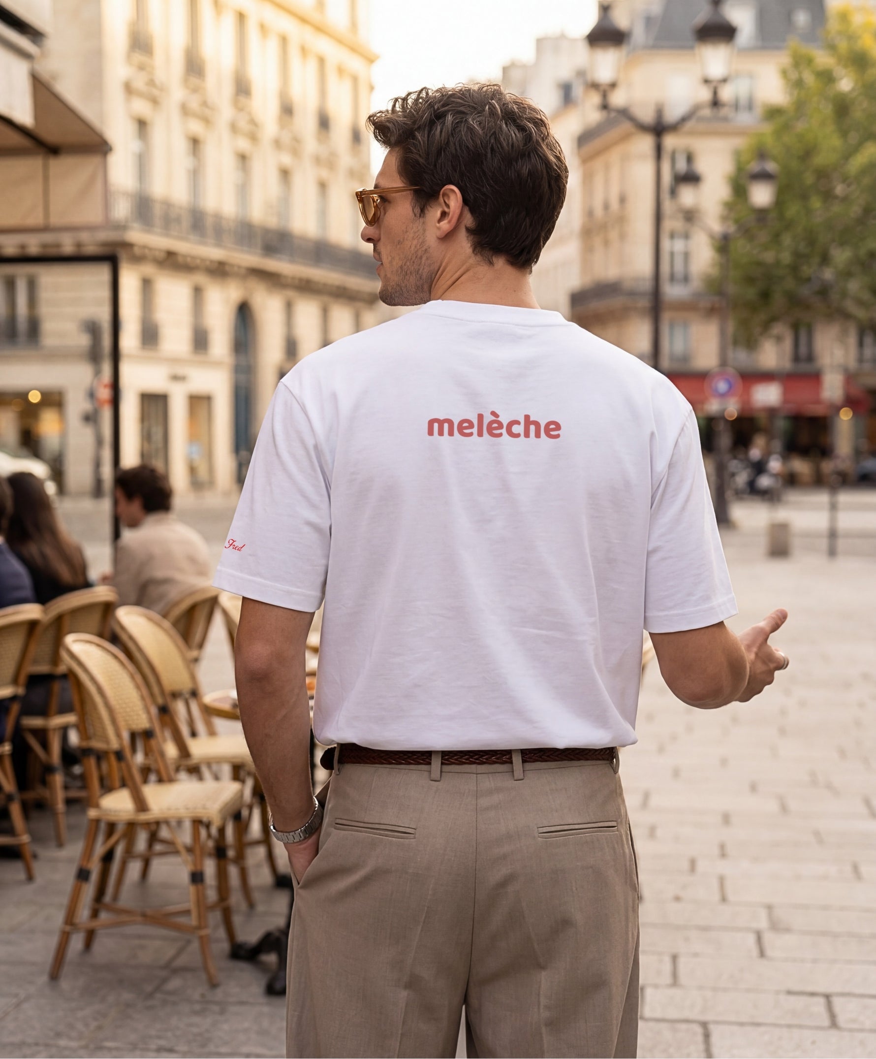 Homme portant le t-shirt oversize blanc « Melon Melèche » avec texte imprimé « melèche » centré dans le dos et logo Salut Fred sur la manche gauche, dans une rue parisienne bordée de cafés et d’immeubles haussmanniens.