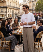 Homme portant le t-shirt oversize blanc « Melon Melèche » avec texte imprimé « melon » centré sur la poitrine et logo Salut Fred sur la manche gauche, dans une terrasse de café parisienne animée entourée d’amis.