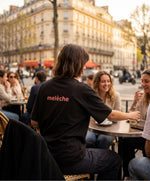 Vue de dos d’une femme portant le t-shirt oversize noir « Melon Melèche » avec texte imprimé « melèche » centré dans le haut du dos, en terrasse parisienne au coucher du soleil.