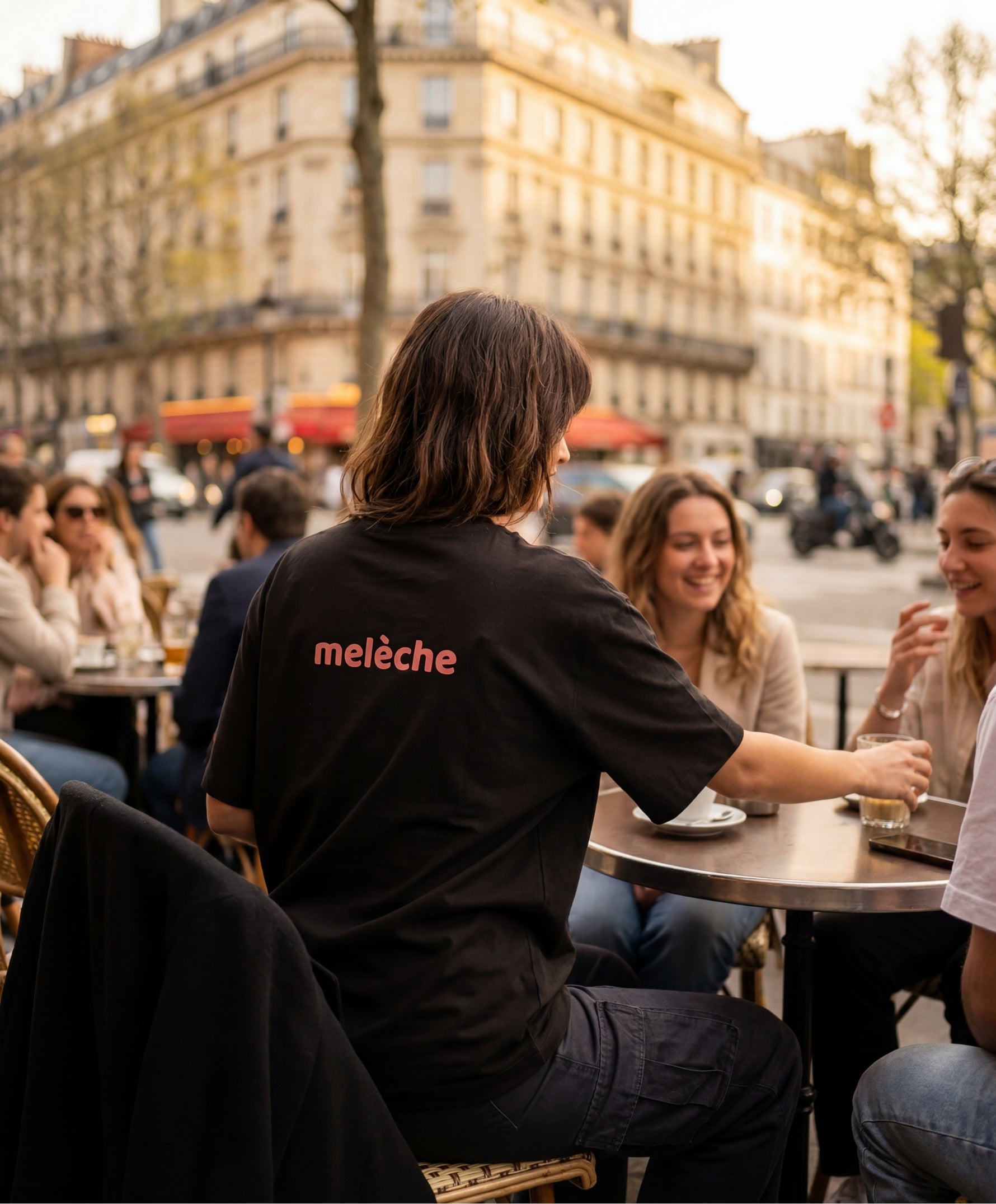 Vue de dos d’une femme portant le t-shirt oversize noir « Melon Melèche » avec texte imprimé « melèche » centré dans le haut du dos, en terrasse parisienne au coucher du soleil.