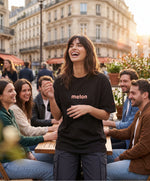 Vue de face d’une femme portant le t-shirt oversize noir « Melon Melèche » avec texte imprimé « melon » centré sur la poitrine et logo Salut Fred sur la manche gauche, attablée en terrasse parisienne au coucher du soleil.