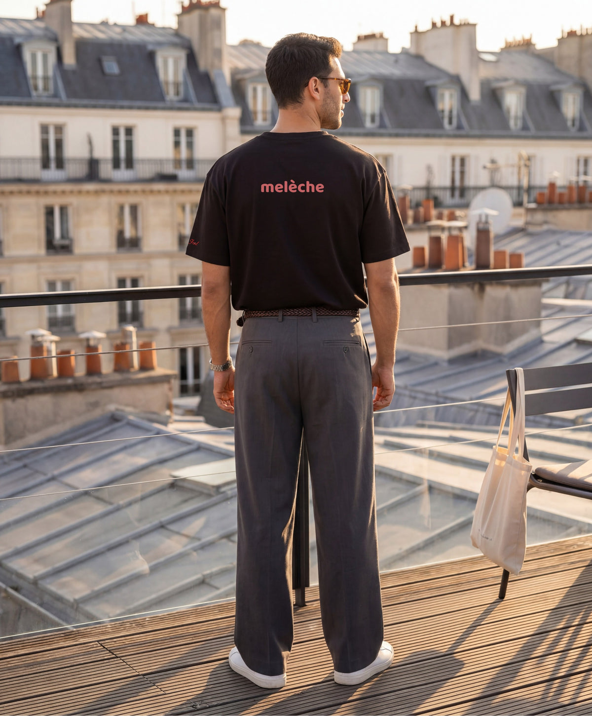Vue de dos d’un homme portant le t-shirt oversize noir « Melon Melèche » avec texte imprimé « melèche » centré dans le haut du dos et logo Salut Fred sur la manche gauche, sur un rooftop parisien au coucher du soleil.