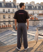 Vue de dos d’un homme portant le t-shirt oversize noir « Melon Melèche » avec texte imprimé « melèche » centré dans le haut du dos et logo Salut Fred sur la manche gauche, sur un rooftop parisien au coucher du soleil.